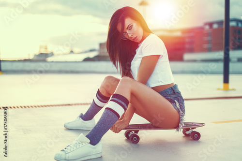 Sexy trendy brunette woman sitting on skateboard in sunset
