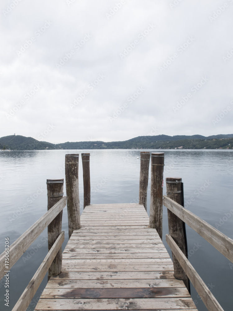 central view of an old wooden pier on the lake d'orta, Italy. Very quiet waters, mountainous background profile, cloudy sky.