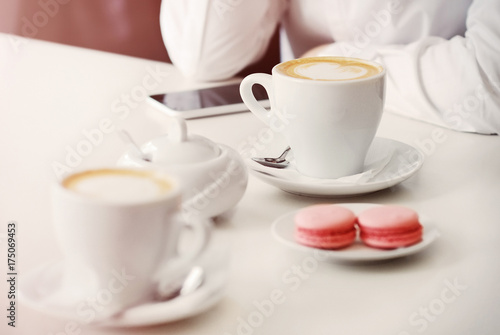 Coffee time. A girl is sitting in a cafe at a table. A coffee cup with coffee, macaroons on a saucer, a sugar bowl and a telephone lie on a white table. Morning in the coffee house