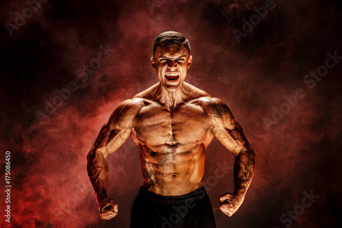 Bodybuilder posing. Fitness tattooed muscled man on red smoke background. Roaring for motivation.