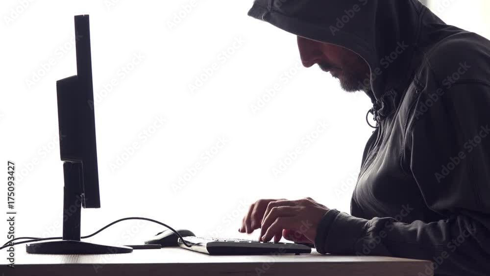 Hooded computer hacker hacking network. Male with black hoodie using computer for identity theft or other criminal online activity.