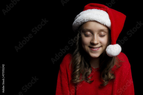 Merry Christmas. A young girl in a red sweater and a Santa Claus hat looks at the viewer.