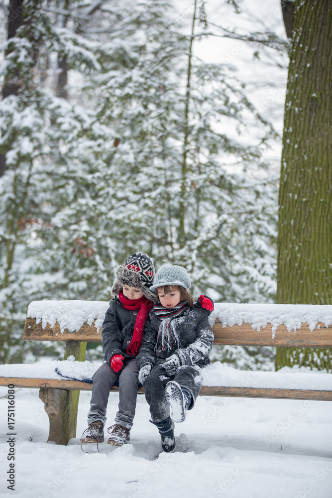 Two children, boy brothers, sitting on a bench in park, wintertime while snowing