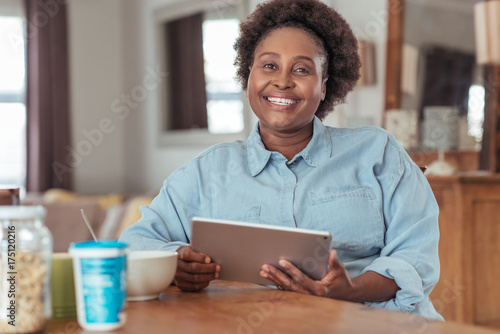 Smiling woman eating breakfast while using a tablet at home