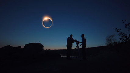 Black silhouettes of people looking through telescope on sun eclipse on shore.