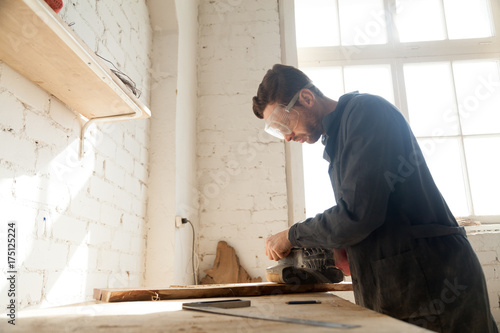 Принти на полотні Carpenter in protective glasses polishes wooden board with handle belt sanding machine on workbench in workshop