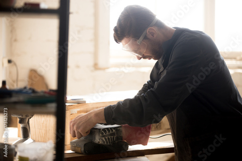 Side view of young joiner wearing protective glasses for safety sanding planks, skilled workman carpenter woodworking in small workshop with equipment tools, building and repairing wooden structures. photo