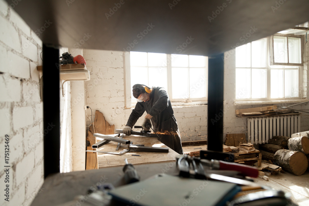 Trained young workman grinding metal parts of construction on table in workshop interior with tools equipment. Manual worker wearing protective clothes working with angle grinder in manufactory Trained young workman grinding metal parts of construction on table in workshop interior with tools equipment. Manual worker wearing protective clothes working with angle grinder in manufactory