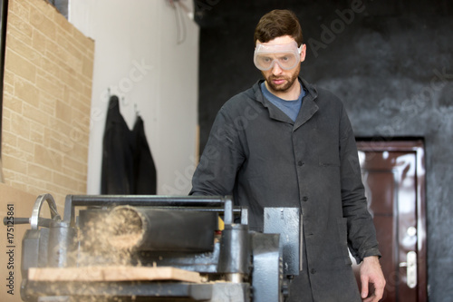 Carpenter in safety glasses standing near machine tool throwing out wooden sawdust while reducing thickness of hardwood board. Small business owner, furniture manufacturer, woodworking industry worker