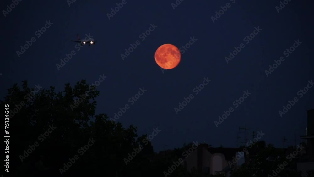 Airplane flies past orange moon in night sky, wide Airplane flies past orange moon in night sky, wide