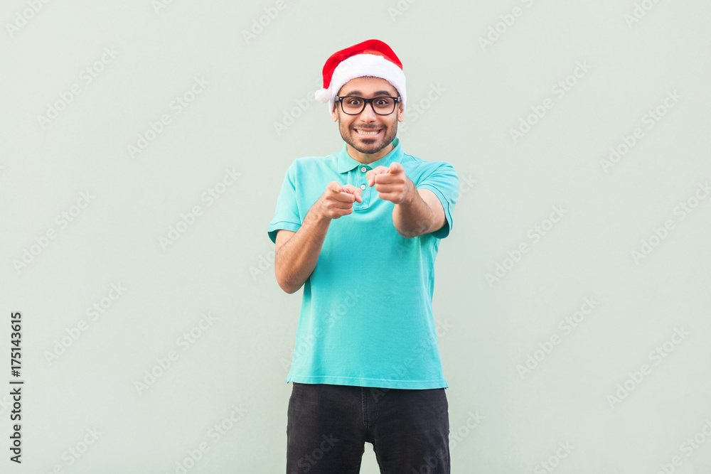 Hey you! Its christmas man! Bearded man in santa hat and glasses, pointing finger and looking at camera with toothy smile.