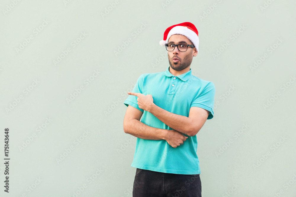 Amazed bearded man in christmas cup with beard in shoked. Pointing away while standing isolated on gray background. Looking at camera with big eyes and open mouth Amazed bearded man in christmas cup with beard in shoked. Pointing away while standing isolated on gray background. Looking at camera with big eyes and open mouth