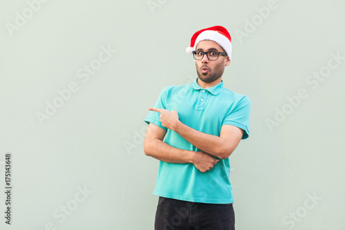 Amazed bearded man in christmas cup with beard in shoked. Pointing away while standing isolated on gray background. Looking at camera with big eyes and open mouth