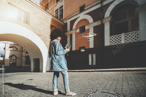 Cheerful young black lady in jean jacket is driving flying drone while standing on pavement stones near architectural arch and holding remote controller for operating quadrupter, sunny autumn day