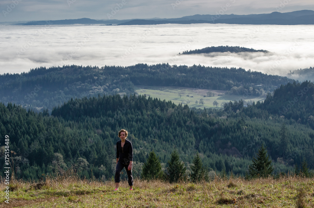 A woman hikes up a hill overlooking a wide valley covered in low clouds.