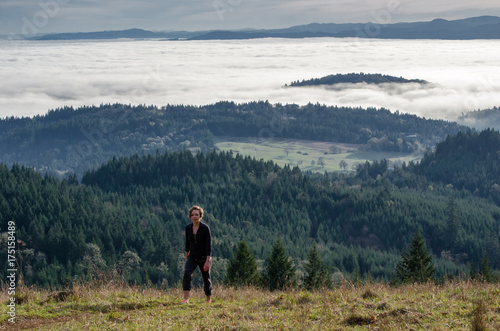 A woman hikes up a hill overlooking a wide valley covered in low clouds.