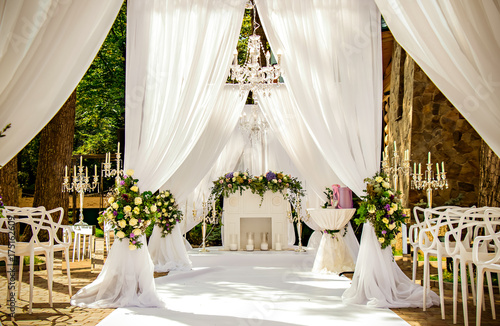 Place for wedding ceremony in white color ,with white fireplace and chandeliers decorated with flowers and white cloth and wooden chairs for guests on each side outdoors. photo