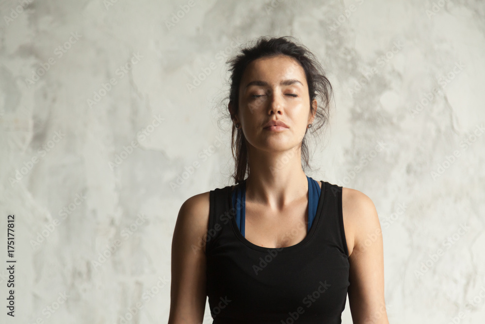 Portrait of young attractive yogi woman with her eyes closed in meditating pose, relaxation exercise, working out, wearing sportswear, black top, indoor close up image, wall background Portrait of young attractive yogi woman with her eyes closed in meditating pose, relaxation exercise, working out, wearing sportswear, black top, indoor close up image, wall background