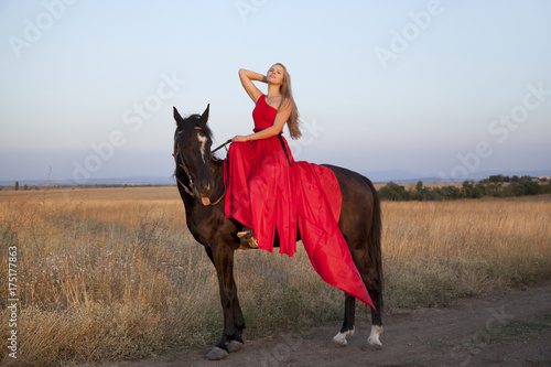 Young woman in a red dress riding a horse. Blonde sitting in the saddle posing in the outdoors