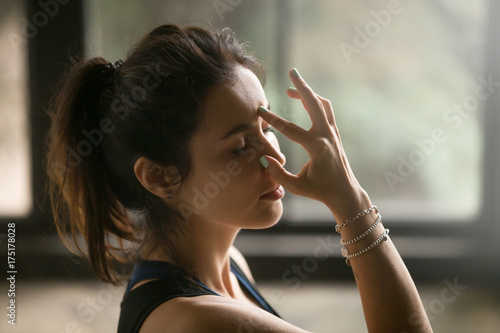 Young attractive woman practicing yoga at home, making Alternate Nostril Breathing exercise, nadi shodhana pranayama pose, working out wearing wrist bracelets, indoor close up image, studio background