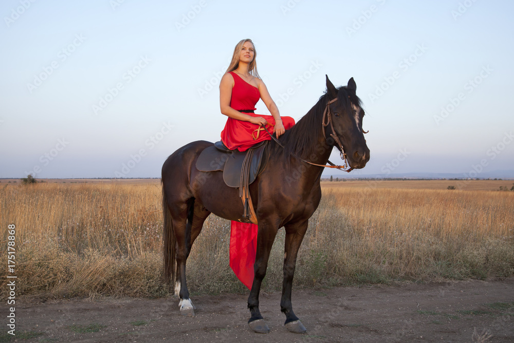 Slim blonde in a long red dress on horseback. Young woman looking into the distance enjoying an evening walk Slim blonde in a long red dress on horseback. Young woman looking into the distance enjoying an evening walk
