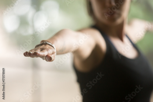 Young sporty woman practicing yoga at home, standing in Warrior Two exercise, Virabhadrasana II pose, working out, wearing sportswear, black top, indoor closeup, studio background, focus on hand