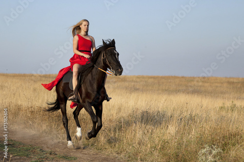 Blonde in a red dress riding a horse. Woman galloping in field on dirt road © vera7388
