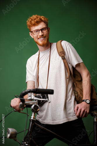 Young cheerful readhead bearded hipster with backpack and retro camera, standing on bicycle