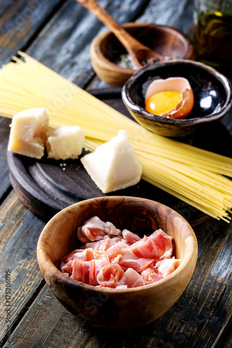 Ingredients for traditional italian pasta alla carbonara. Uncooked spaghetti, pancetta bacon, parmesan cheese, egg yolk, salt, pepper in olive wood bowls over old plank background.
