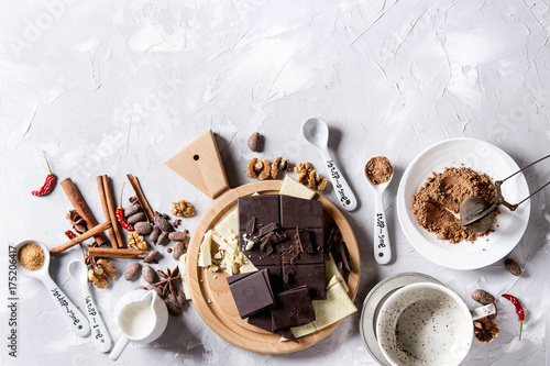 Ingredients for cooking hot chocolate. White and dark chopping chocolate on board, cocoa powder, cocoa beans, cream, cinnamon, sugar in spoons. Over gray texture background. Top view with copy space