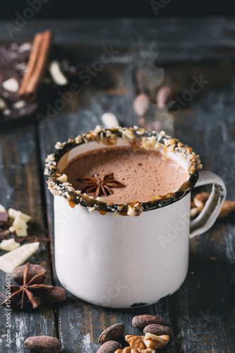 Vintage mug of hot chocolate, decor with nuts, caramel, spices. Ingredients above. Chopped dark and white chocolate, cocoa beans, anise over old wooden table. Dark rustic style