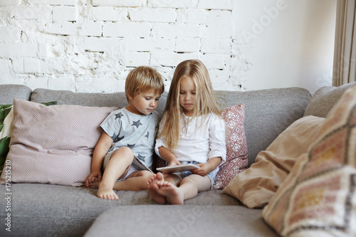 Two siblings using digital tablet together, sitting on couch in living room, watching cartoons online via social media. Little boy watching his elder sister playing video games on touchpad computer