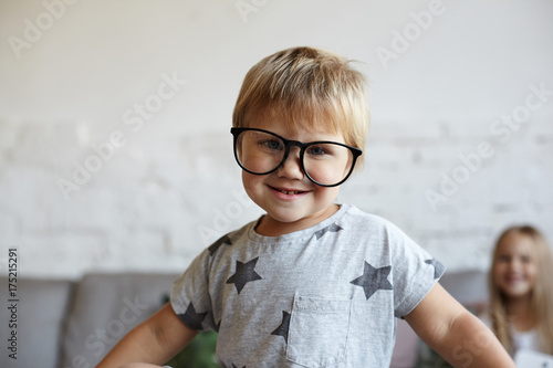 Close up of adorable cute little boy wearing t-shirt with stars and big trendy eyeglasses in black frame, looking and smiling at camera with his sister sitting on couch in background. Selective focus
