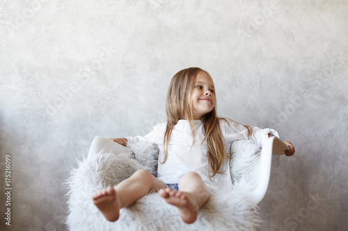 Happy childhood concept. Isolated shot of adorable European little girl relaxing barefooted on white comfortable chair and looking sideways with positive cute smile, posing at blank grey wall