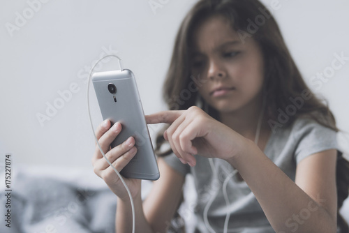 A little black-haired girl in white headphones with a gray smartphone in her hands switches music