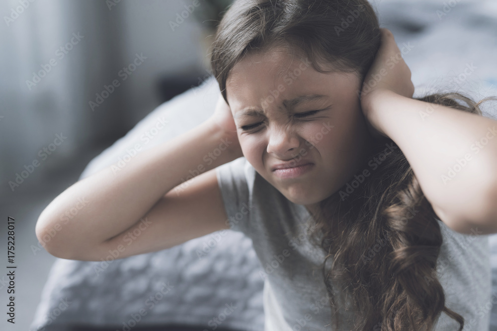 The black-haired girl sits next to the bed frowning and covering her ears with her hands The black-haired girl sits next to the bed frowning and covering her ears with her hands