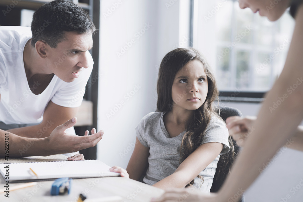 Obraz premium A man and a woman scold a little girl who sits with a desk. The girl angrily looks at the woman