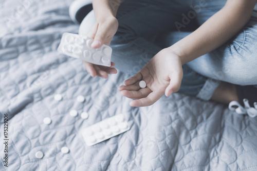 A teenage girl sits on a bed and holds an opened pack of pills in her hands, some of which are lying near her
