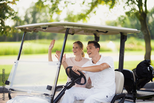 A man and a woman are sitting in a white golf cart and waving their hands to someone ahead of them © VadimGuzhva