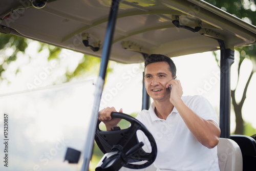 A man is driving a golf car and talking to someone on a smartphone
