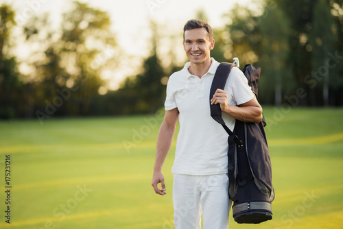 A man in a white suit walks around the golf course with a golf club bag and smiles