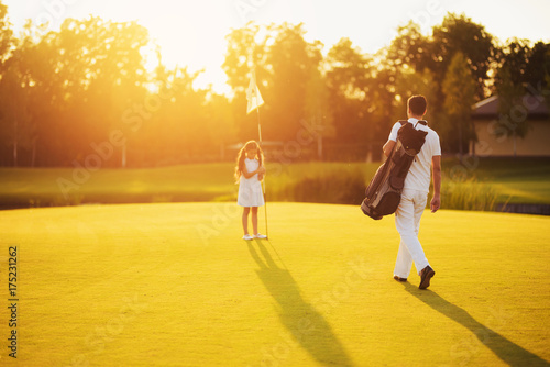 A man in a white suit and a golf club bag goes towards the girl who stands next to the hole in the sunset