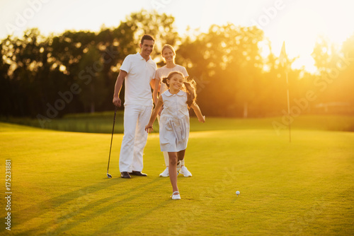 Happy family posing on a golf course on a sunset background. The girl smiles and runs towards the camera