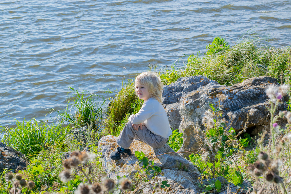 Blond thoughtful boy sitting at a river bank on a rocks - Rusiian Urals wind sunny day