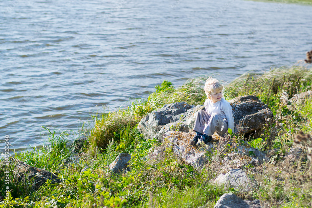 Blond thoughtful boy sitting at a river bank on a rocks - Rusiian Urals wind sunny day