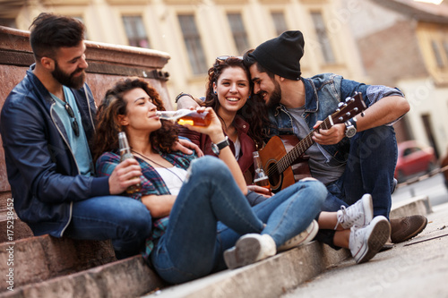 Group of young friends hangout on city square .They playing guitar and sings.