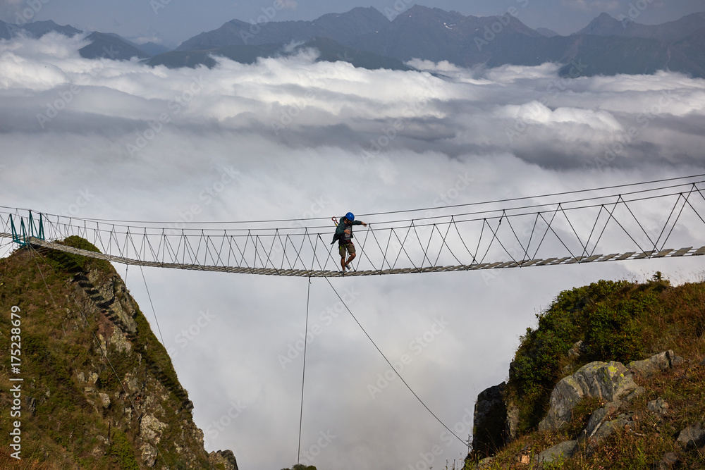 Man walking on suspension bridge and looking at cloudy mountains below.