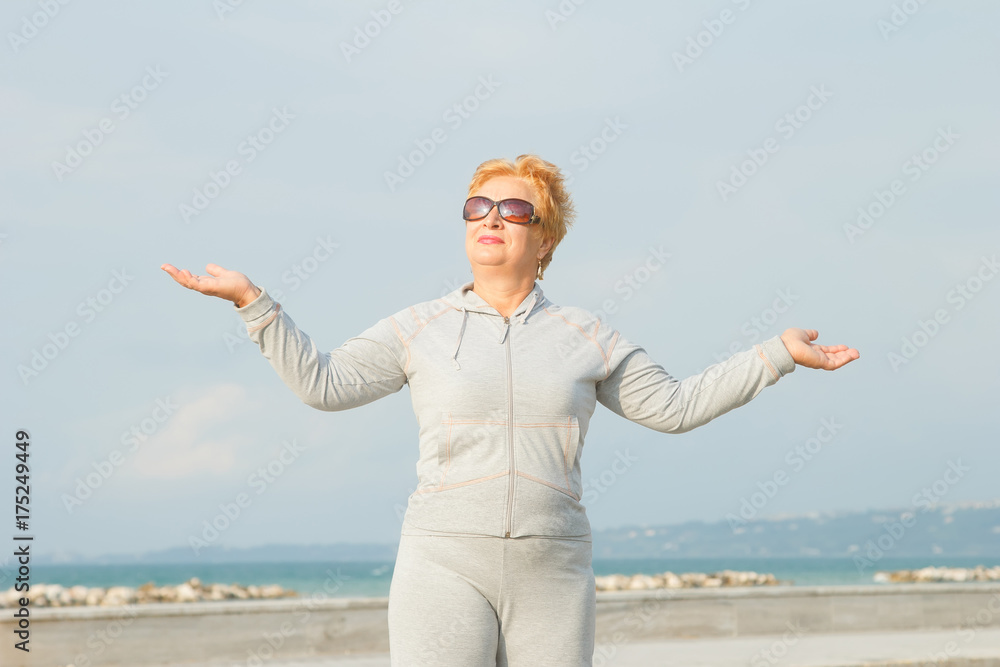 Adult woman on the background of the sea. Senior woman in tracksuit enjoying life. Fitness classes in the open air near the sea and the beach. Lifestyle adult retired. Adult woman on the background of the sea. Senior woman in tracksuit enjoying life. Fitness classes in the open air near the sea and the beach. Lifestyle adult retired.