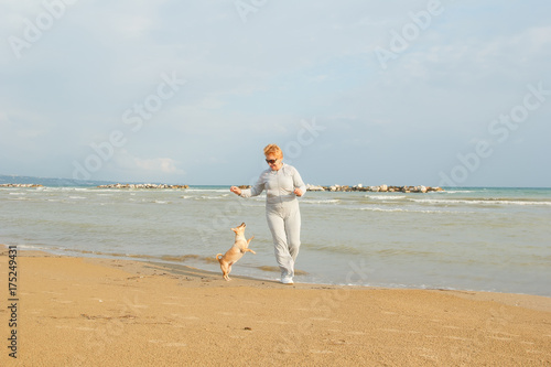 Adult woman on the background of the sea playing with the dog. Senior woman in tracksuit enjoying life. Fitness and walking outdoors near the sea and the beach. Lifestyle adult retired.