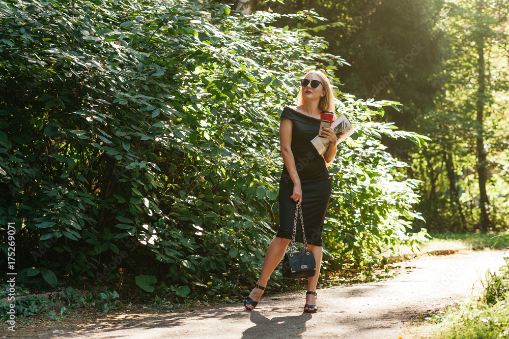 Young business lady in black dress and glasses with coffee and a bag walking in the park